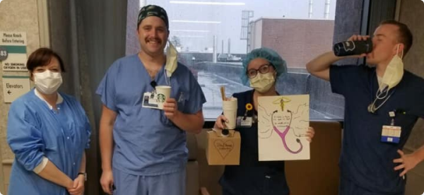 Four healthcare workers in scrubs, wearing masks, posing with coffee and a handmade drawing of a stethoscope.