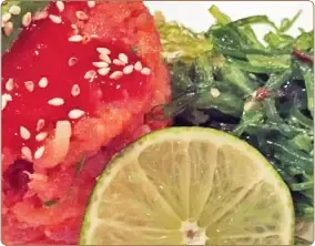 A close-up of a salmon tartare dish topped with red sauce and sesame seeds, served with a lime slice and seaweed salad.
