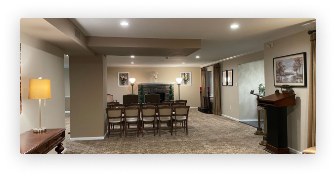 A carpeted room with chairs facing a fireplace, a podium, and soft lighting, likely a funeral home or event space.