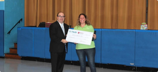 Two people stand in a gym, one in a suit and one in a green shirt, holding a large ceremonial donation check.