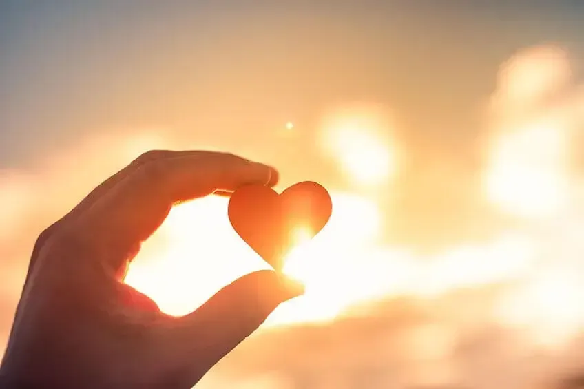 A hand holds a heart-shaped object against a golden sunset, with light glowing through the edges of the heart.