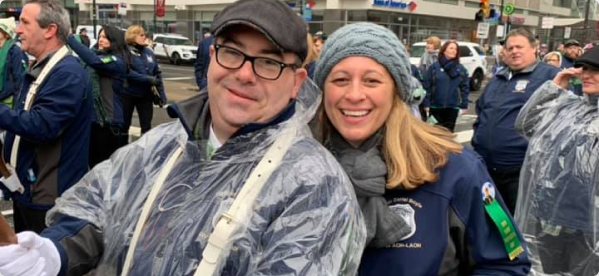 A smiling couple in rain gear and winter hats pose for a photo outdoors during a public event.