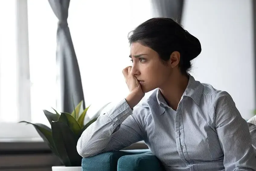 A person rests their chin on their hand, gazing away with a pensive expression while sitting indoors near a window.