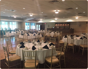 A banquet hall with round tables set with white tablecloths, black napkins, and gold-colored chairs, and a bar in the rear.