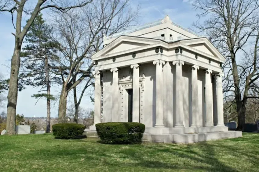 A neoclassical stone mausoleum with columns and a triangular pediment, situated in a park-like cemetery setting.