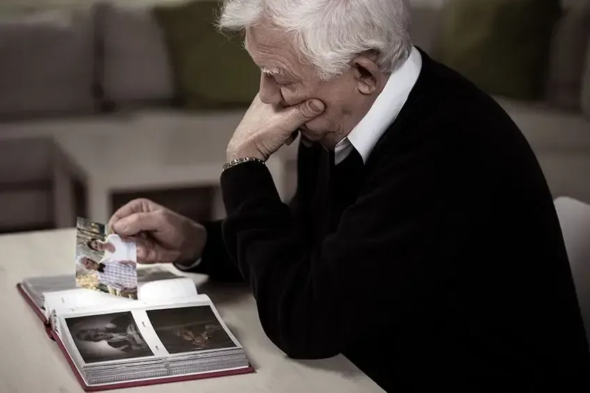 A person sits at a table looking down at an open photo album, holding one photograph while resting their chin on a hand.