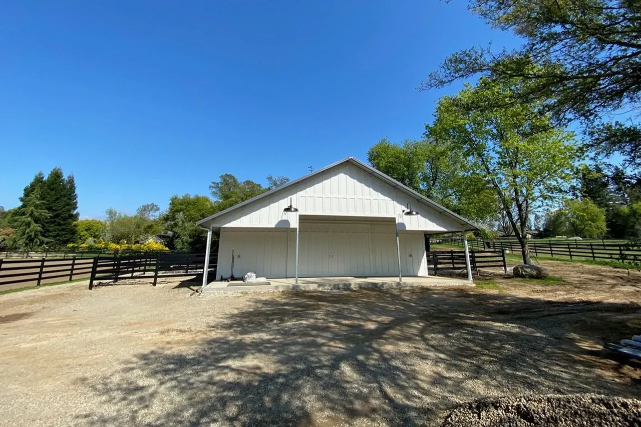 A white barn is sitting in the middle of a dirt field next to a fence.