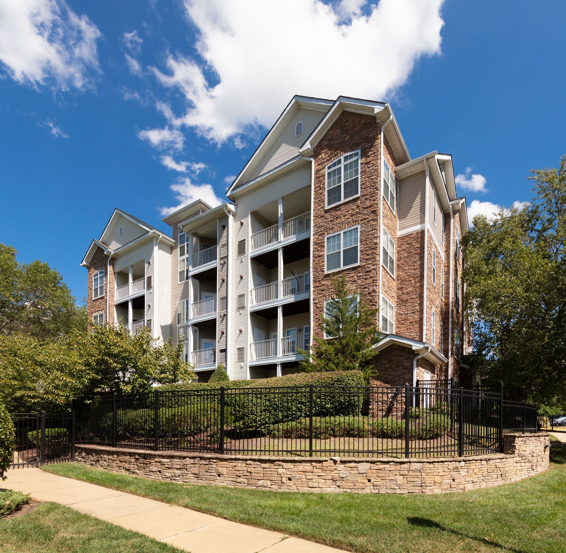 Apartment building with brick and white accents, balconies, and a black fence under a blue sky.