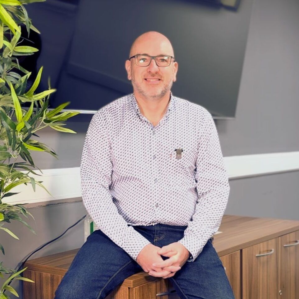A bald man wearing glasses is sitting on a wooden cabinet.