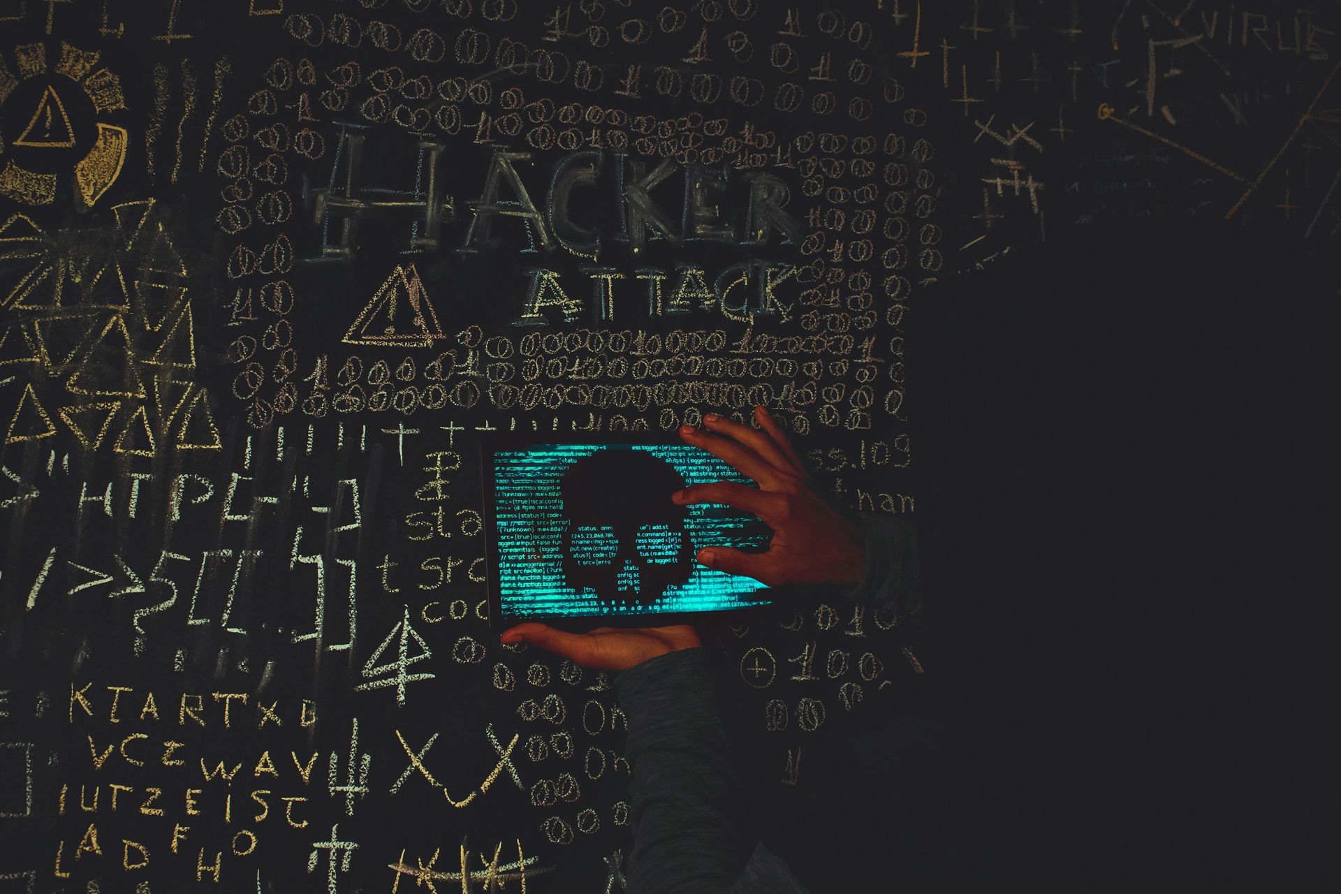 Hands holding a tablet displaying a skull, in front of a wall covered in hacker symbols and the word