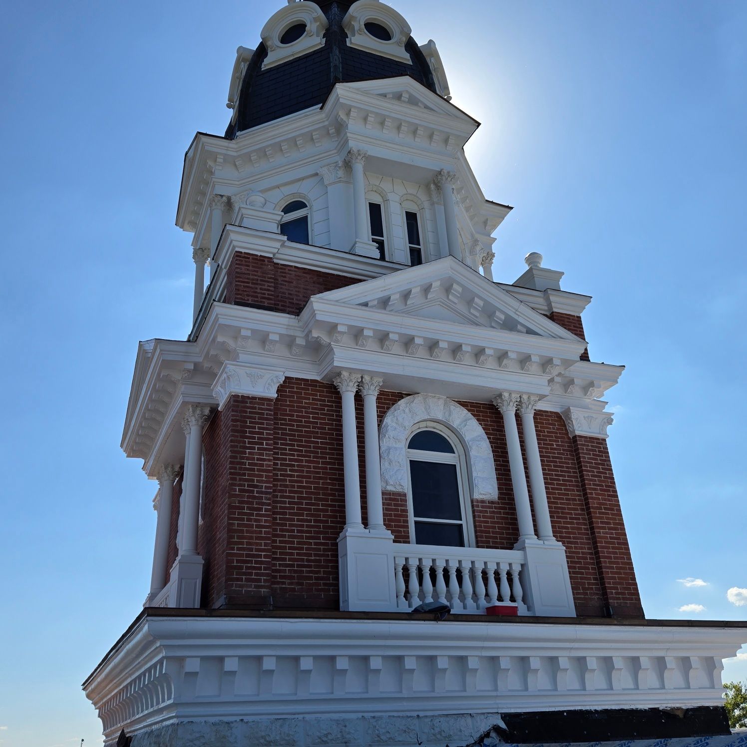 Brick and white tower with arched windows, topped with a black roof, against a blue sky with the sun.