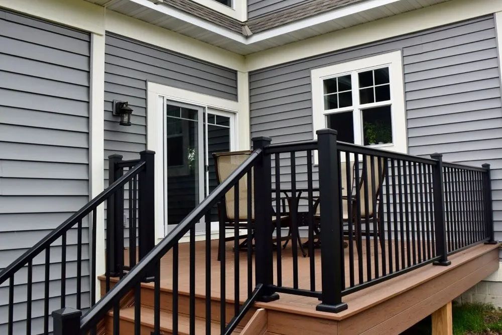 Black railing surrounds a composite deck with sliding glass doors, gray siding, and a window.