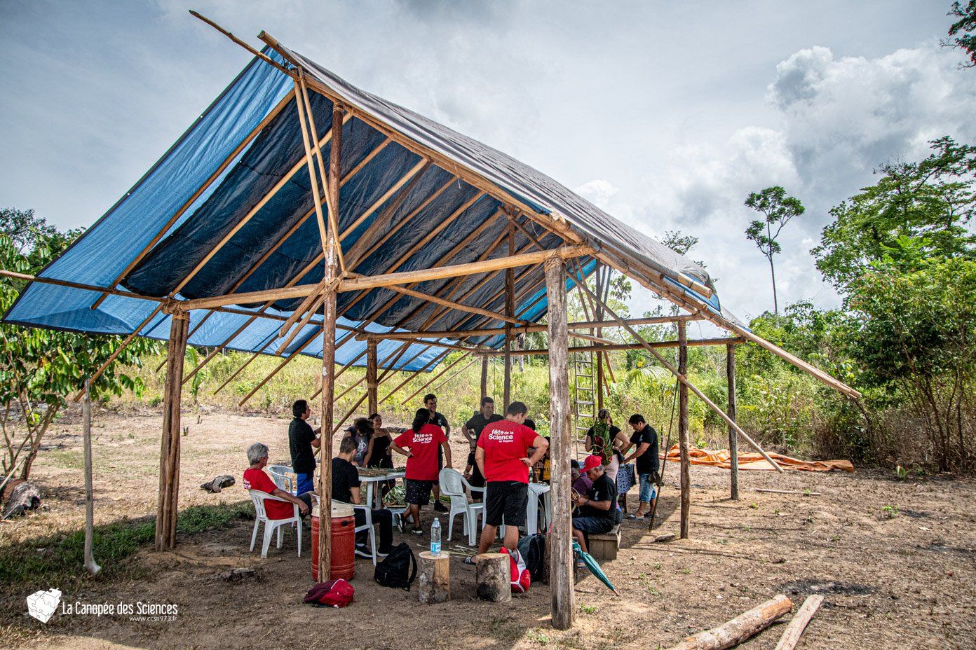 Un groupe de personnes est assis sous une bâche bleue sous une structure en bois.