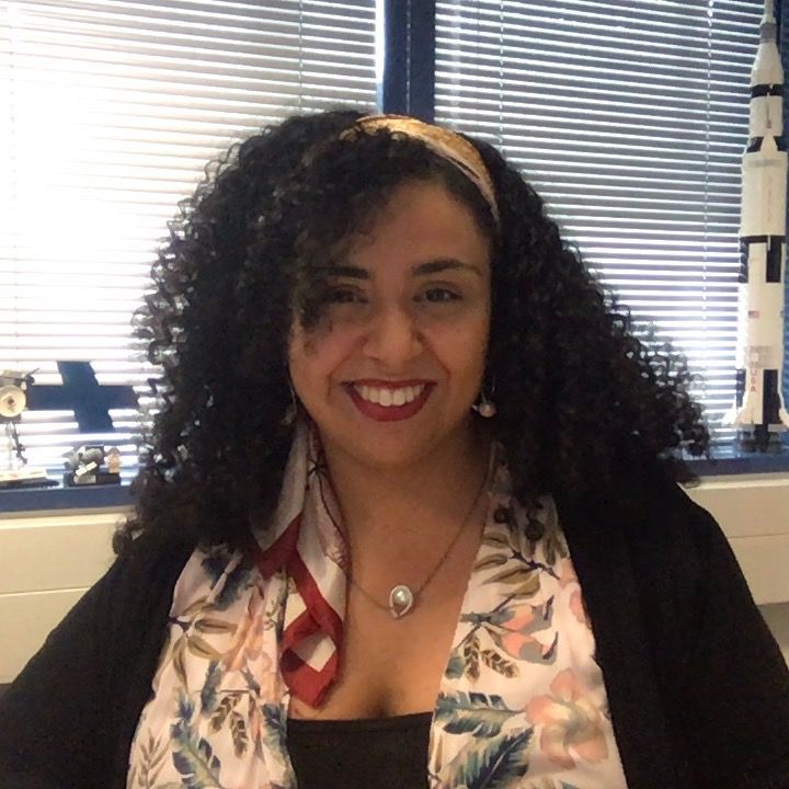 Une femme aux cheveux bouclés sourit devant une fenêtre avec des stores