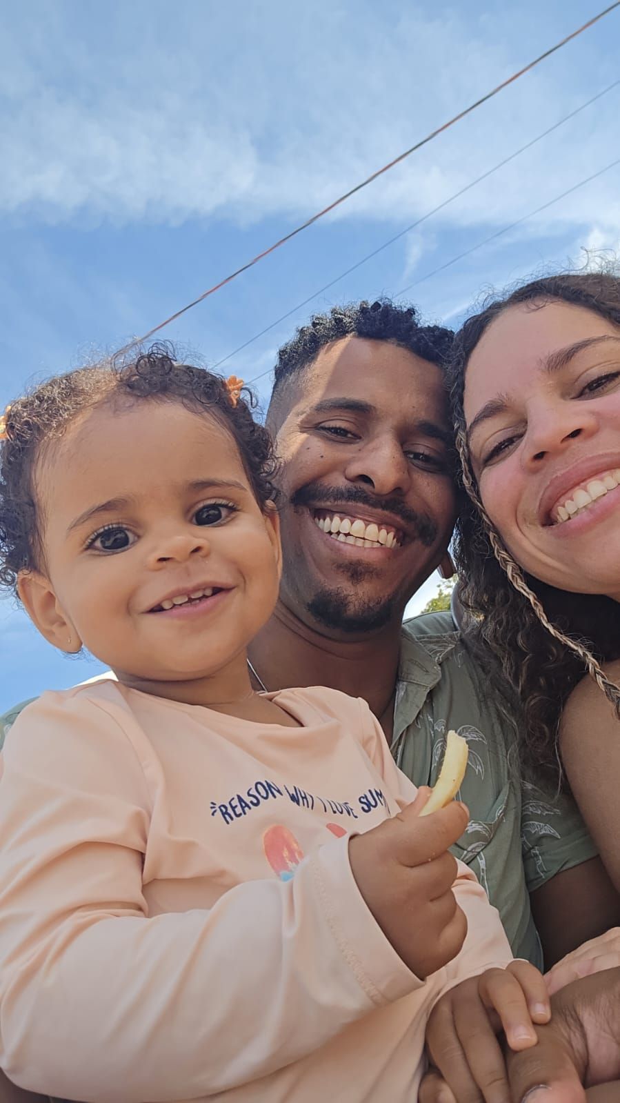 Foto de uma família sorrindo ao ar livre. Um casal e uma criança pequena aparecem em selfie.