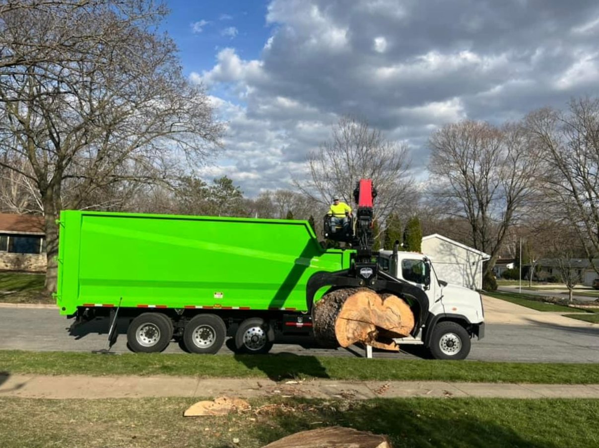 A green garbage truck is driving down a street next to a tree stump.
