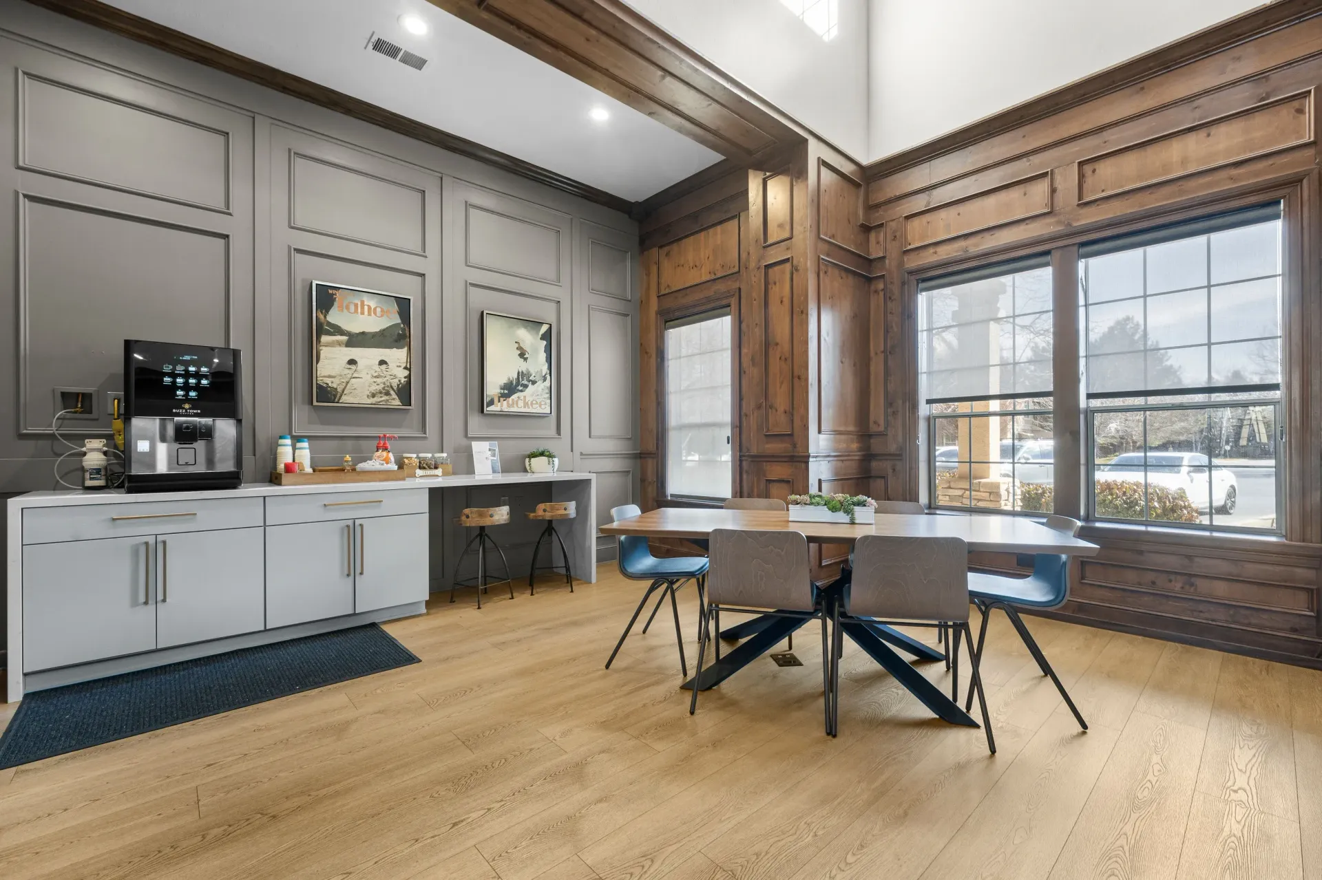 Community lounge with coffee station, counter, and wooden dining table by large window.
