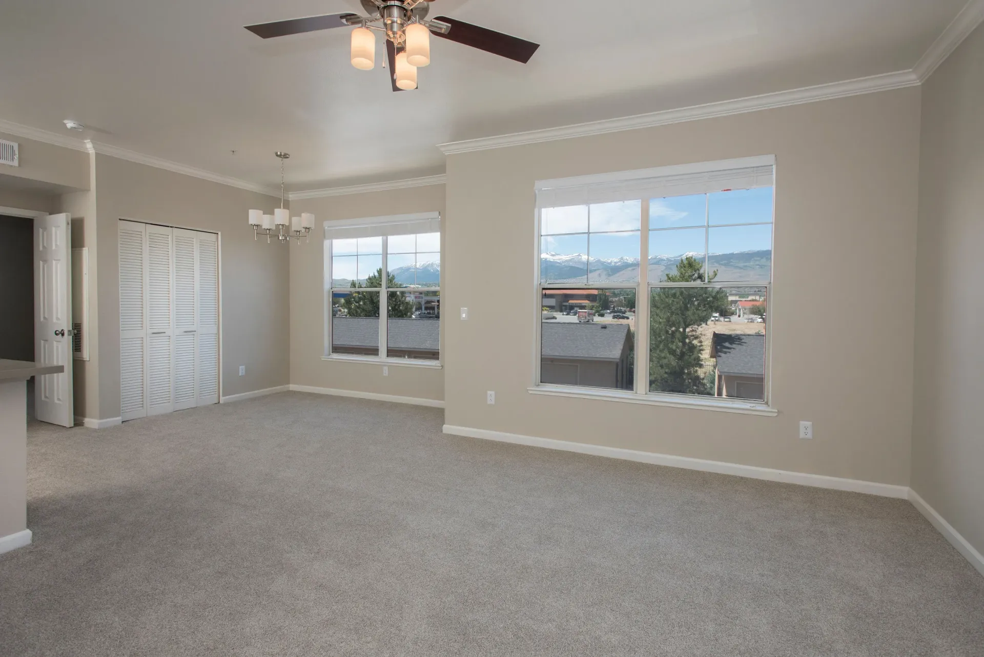 Living room in apartment with beige walls, beige carpet, large windows, and ceiling fan.