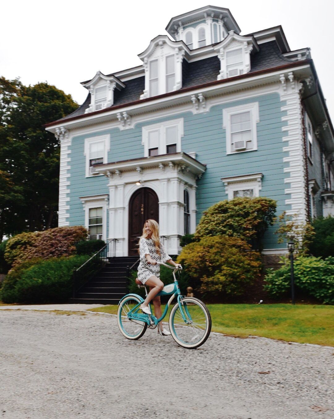 A person in light clothing rides a matching light blue bicycle in front of a historic, pale blue three-story mansion.
