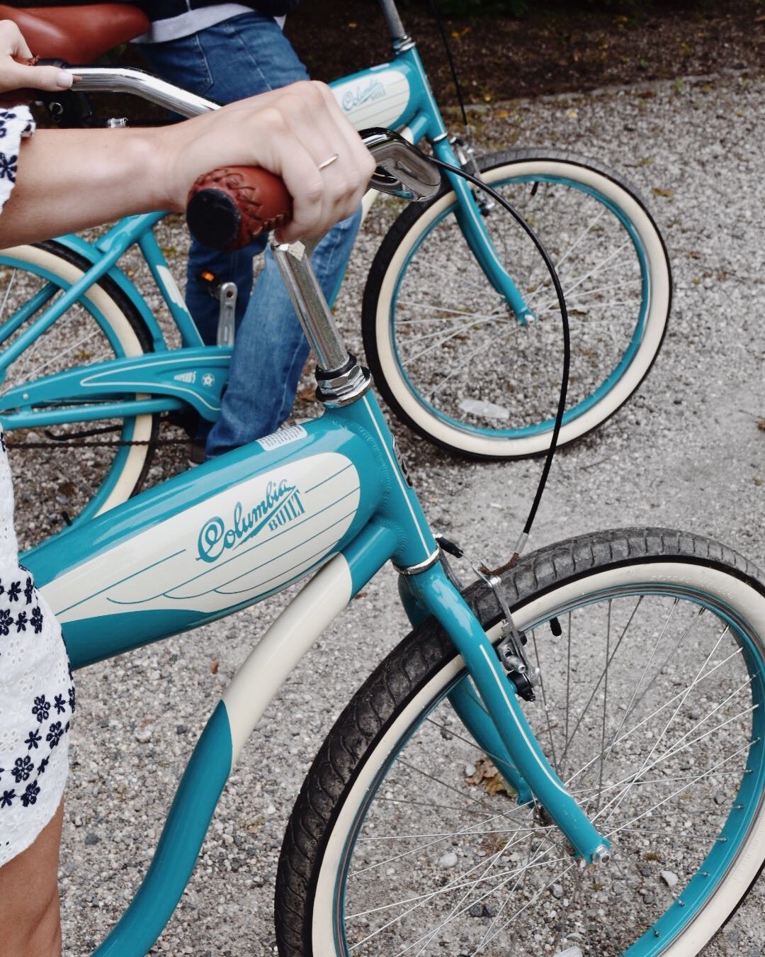 A close-up view of a person’s hand on the handlebar of a turquoise and white cruiser bicycle on a gravel path.