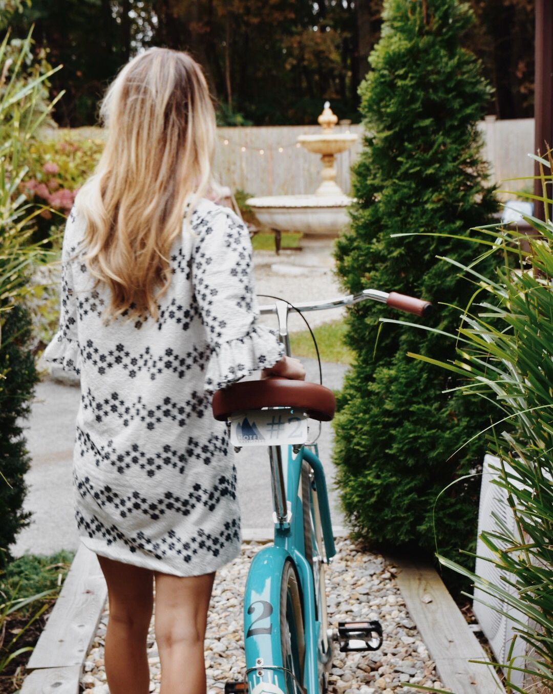 A person in a patterned white dress walks a teal cruiser bicycle on a gravel path toward a stone fountain in a garden.