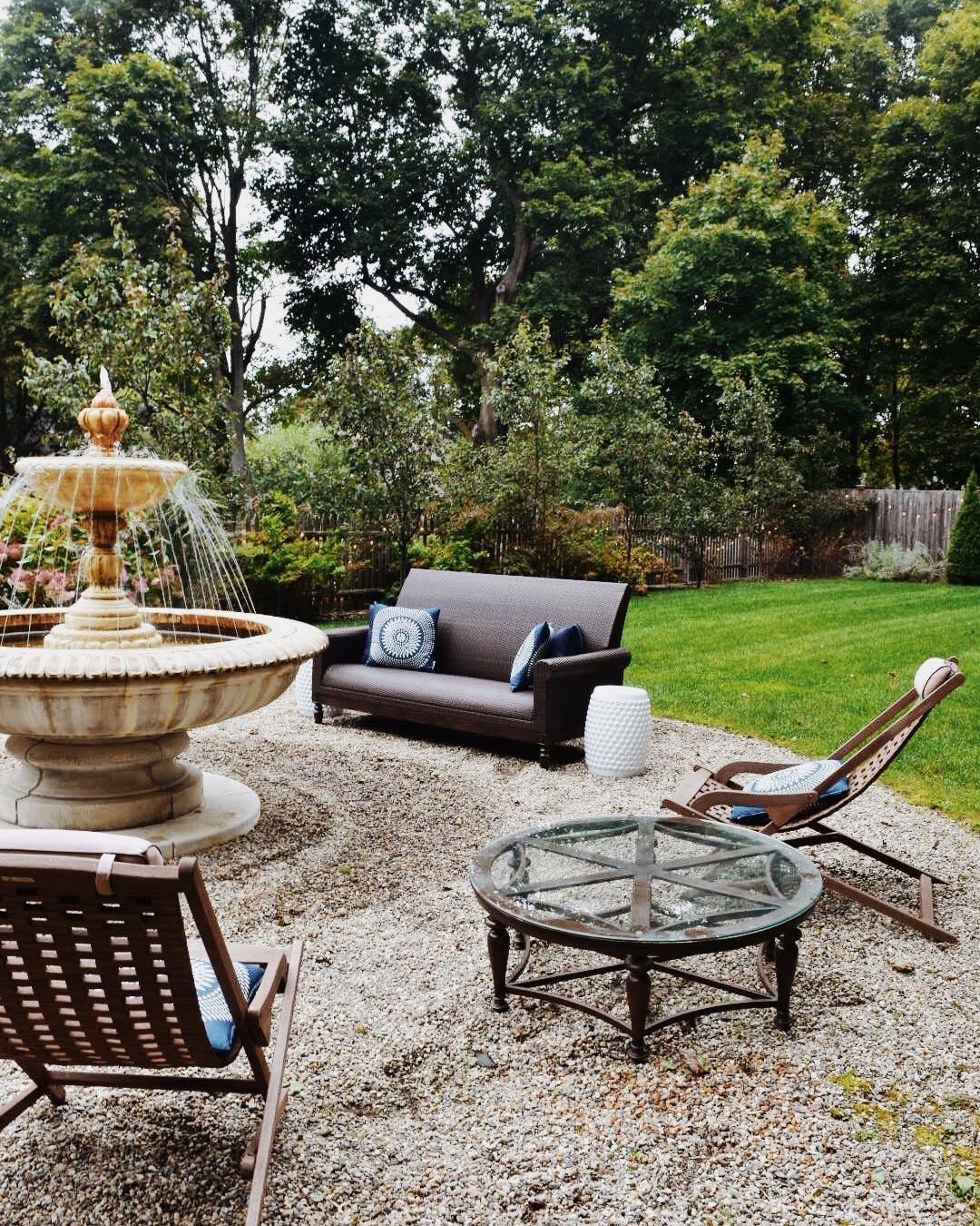 An outdoor seating area with a stone fountain, brown sofa, glass-topped table, and two wooden lounge chairs on gravel.