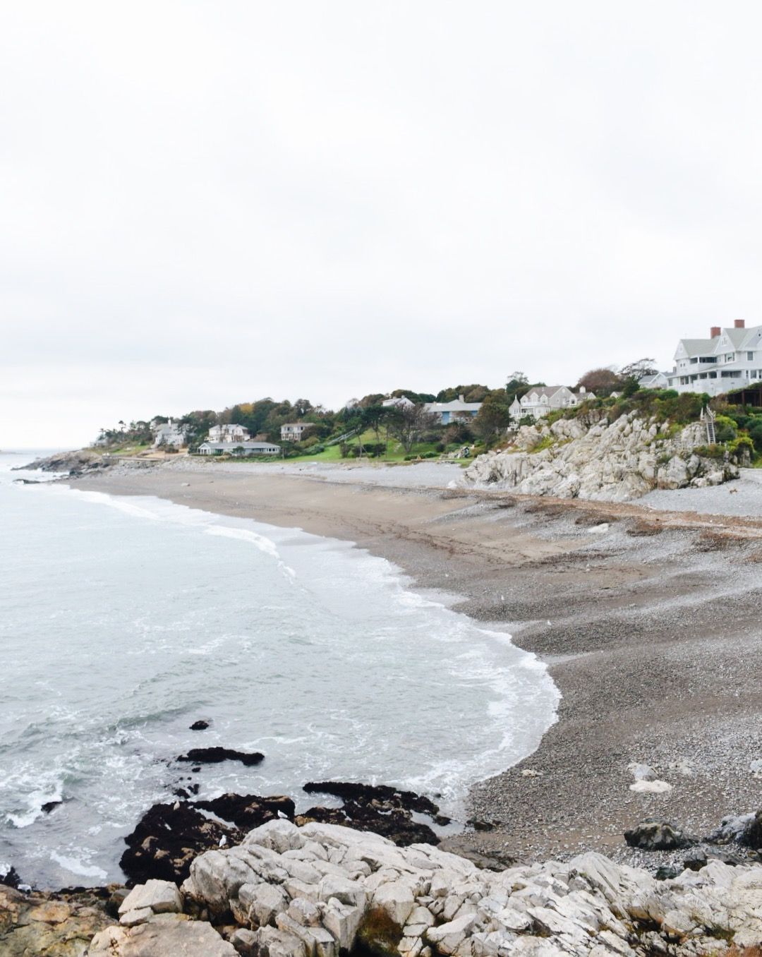 A stony beach curves along a coastline with houses nestled among trees and rocky outcroppings under a cloudy sky.