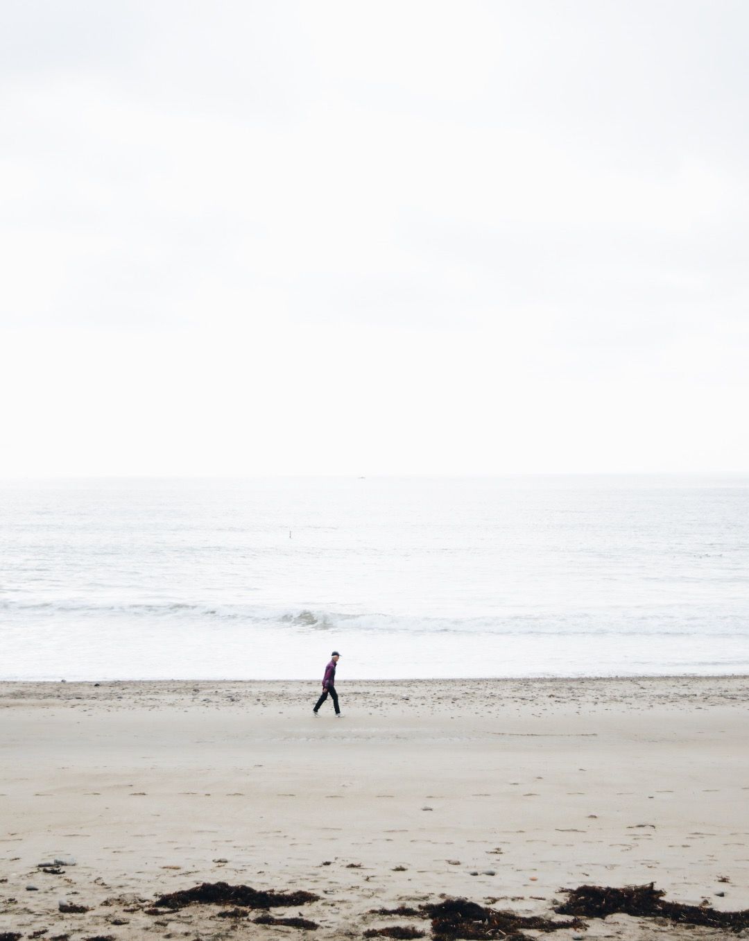 A lone person walks along a wide, pale sandy beach next to the ocean under a bright, overcast sky.