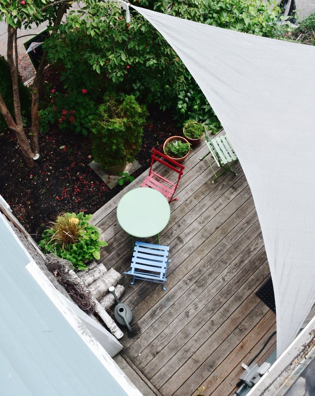 An elevated view of a wooden deck with a small table, colorful chairs, potted plants, and a large triangular shade sail.