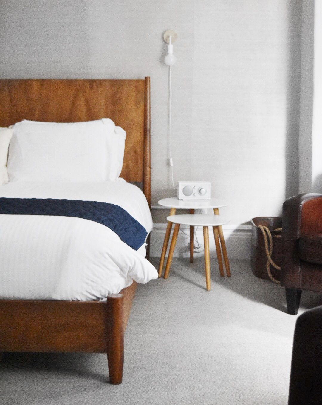 A modern bedroom features a wooden bed with white bedding, nested side tables, and a leather chair on gray carpeting.
