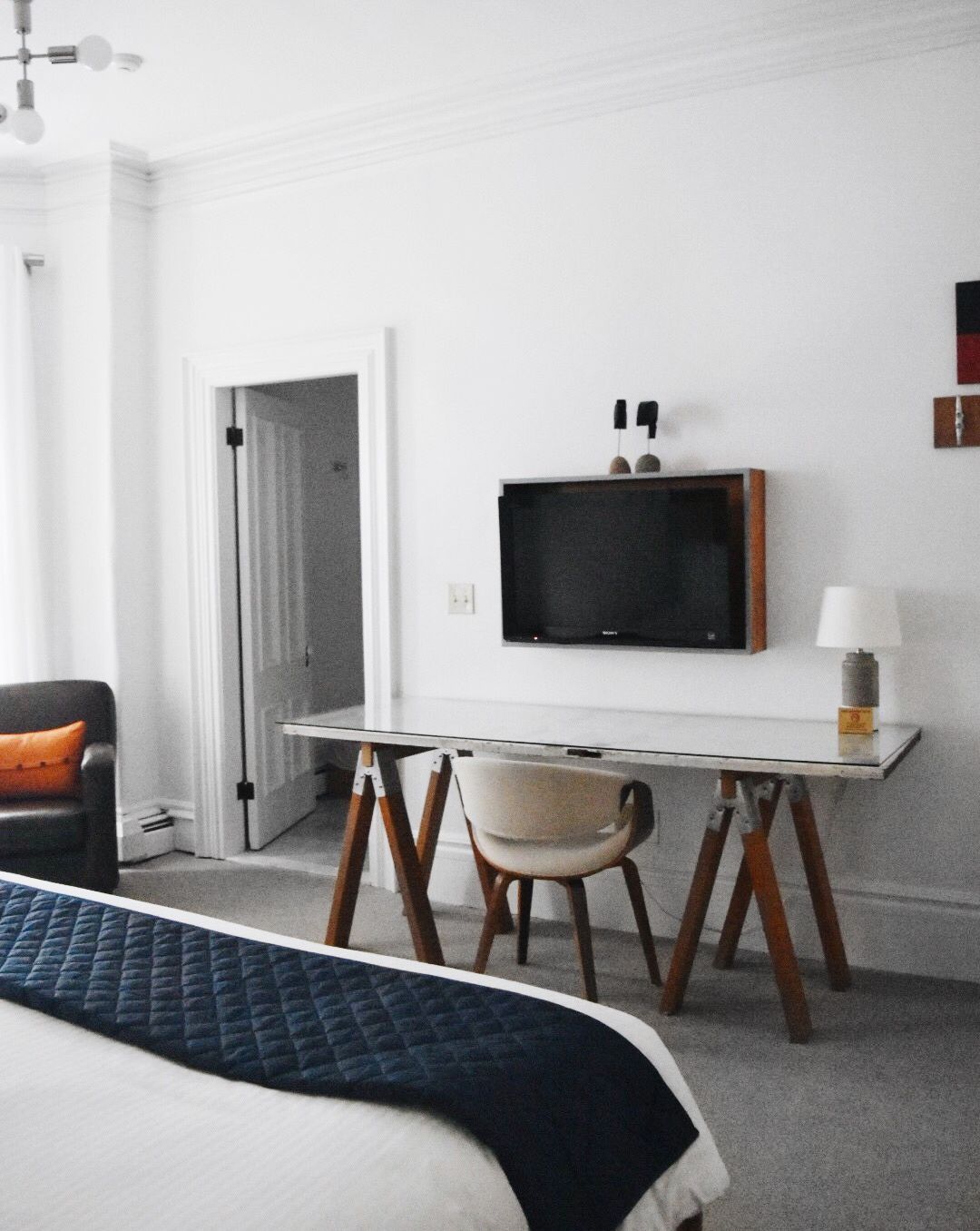 A hotel room with a bed, a desk featuring a sawhorse base, a mounted TV, and an open doorway to a bathroom.