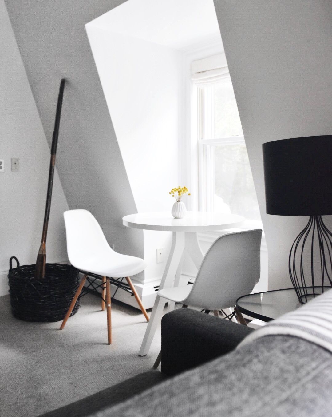 A small white dining table with two white Eames-style chairs by a window in a room with a broom in a basket and a lamp.