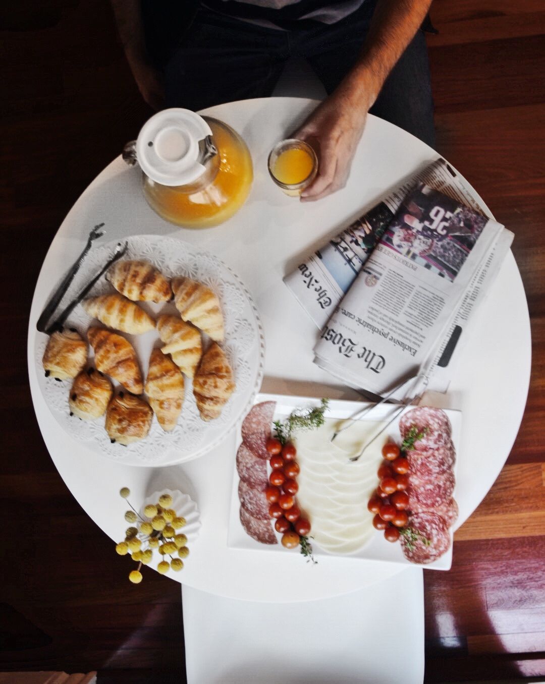 A top-down view of a white table featuring a plate of croissants, a charcuterie platter, orange juice, and a newspaper.