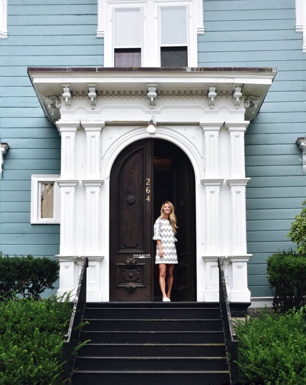 A person stands in the open, dark-wood arched doorway of a blue-sided house with white trim and black front steps.