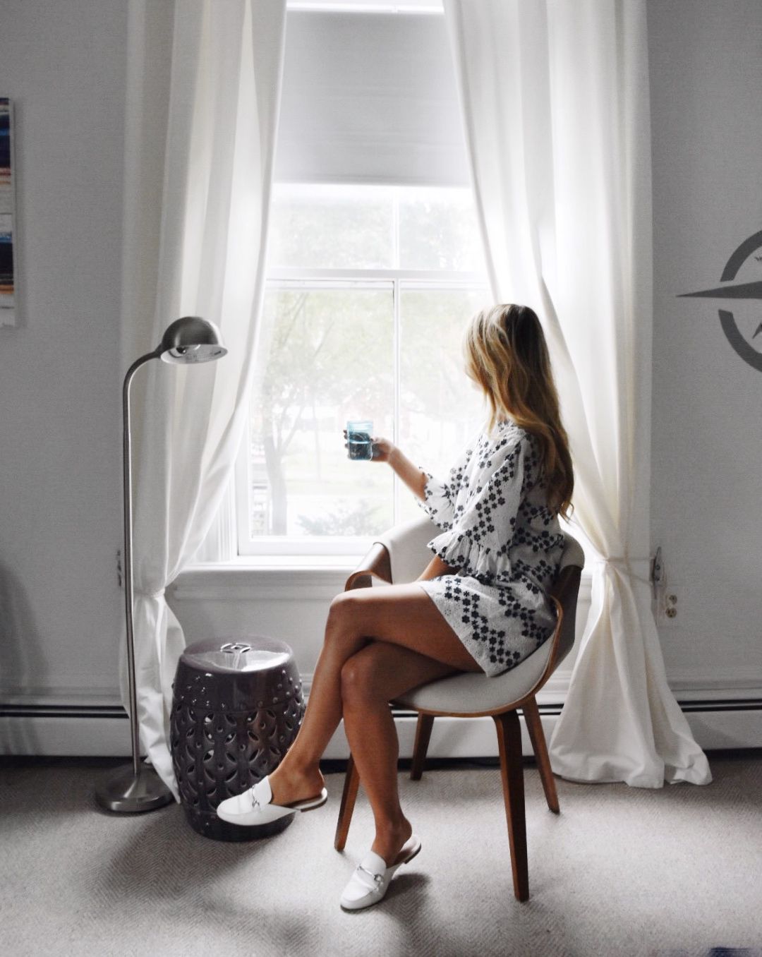A person sits in a chair by a window, holding a glass, wearing a patterned white dress and white slide shoes.