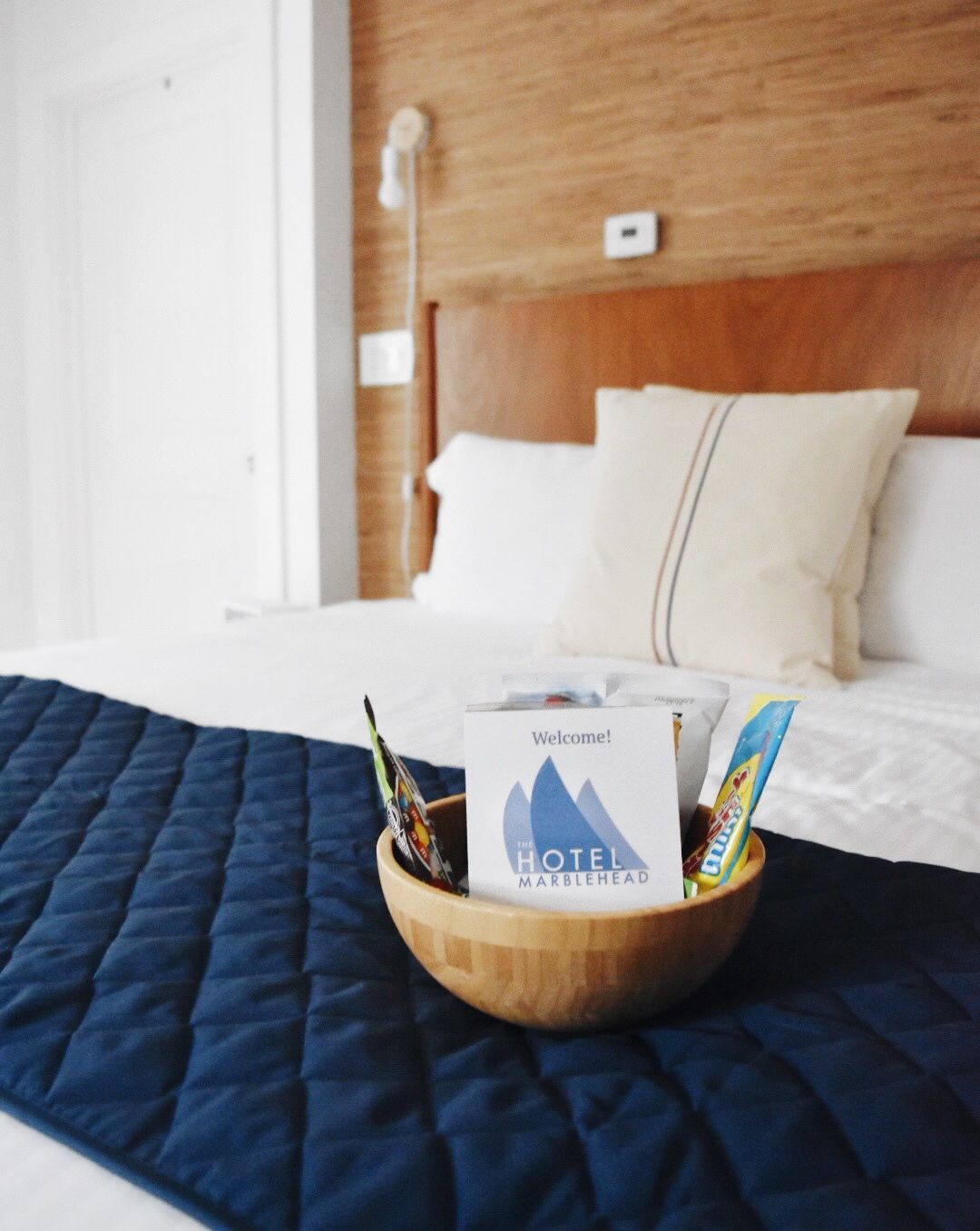 A bowl with snacks and an info card sits on a bed with white linens and a blue quilted runner in a hotel room.