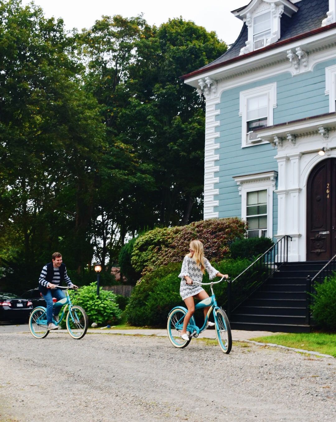Two people ride light blue bicycles along a gravel path in front of a blue house with white trim and a black staircase.