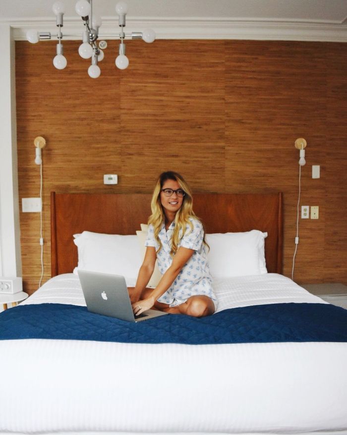 A person with long blonde hair and glasses wearing patterned pajamas sits on a bed using a laptop in a modern bedroom.