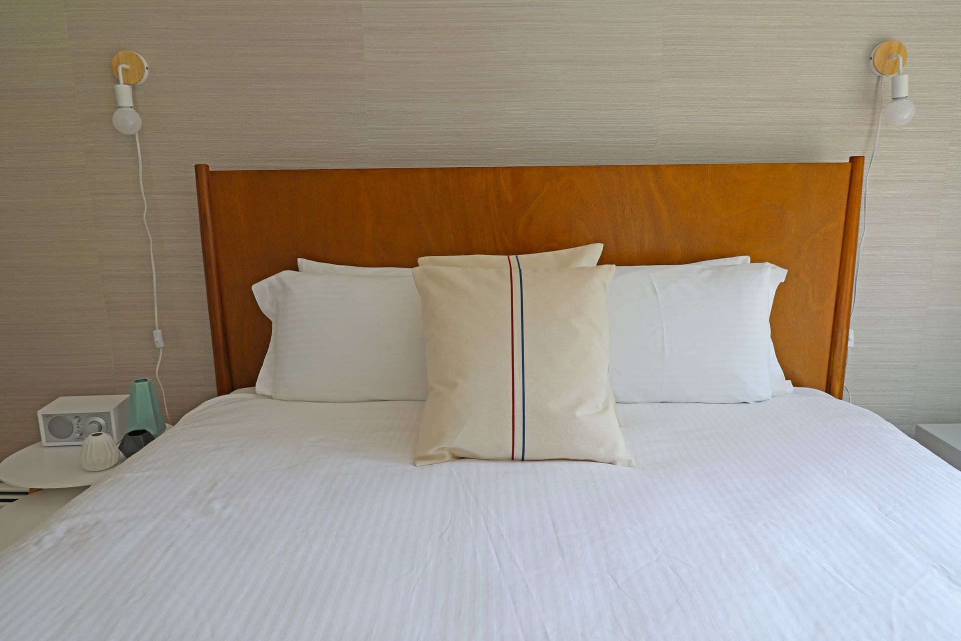 A bright bedroom featuring a queen bed with white linens and a blue runner, light wood furniture, and a television.