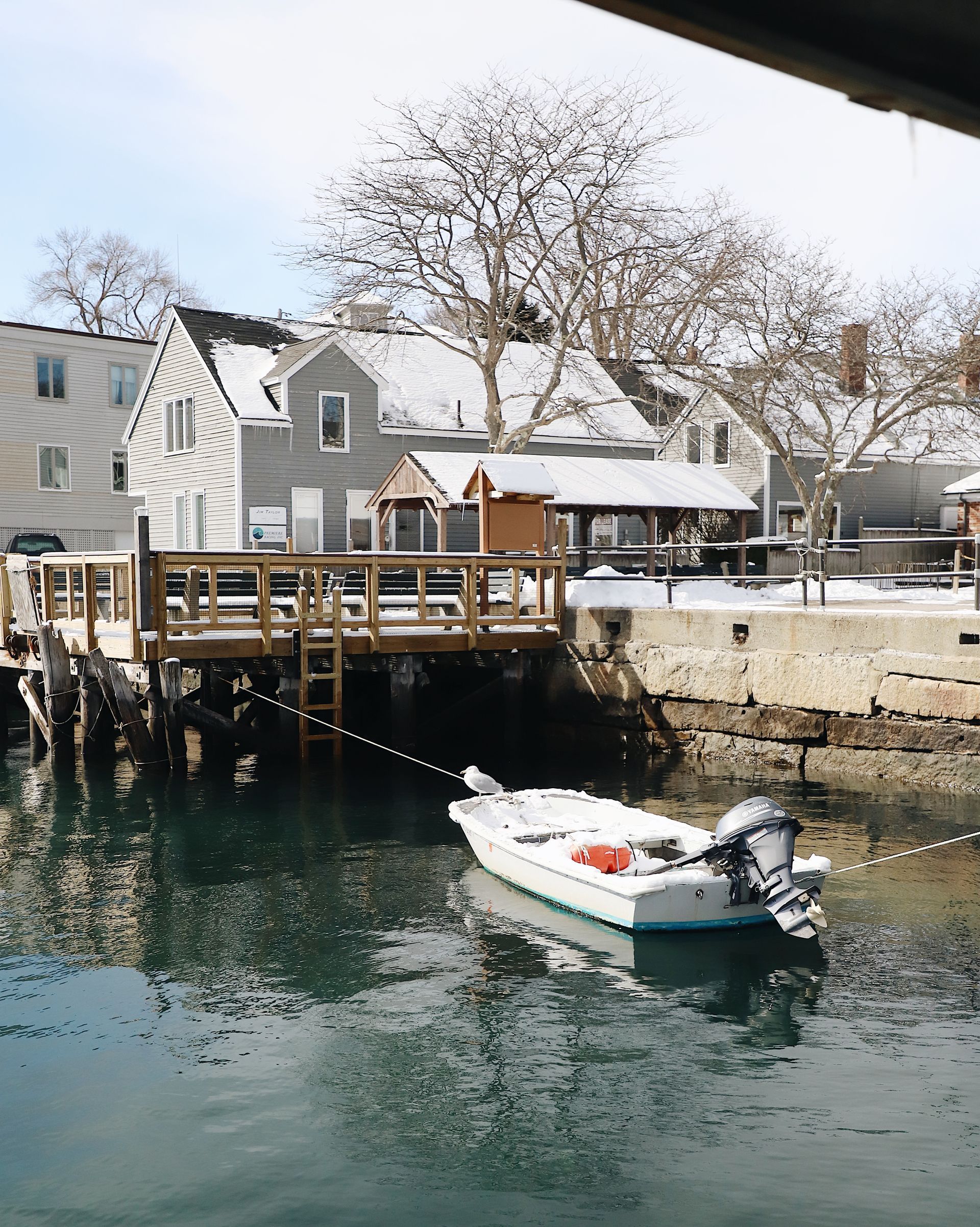 A small white boat with an outboard motor floats on calm water in front of a snow-covered house and wooden dock.