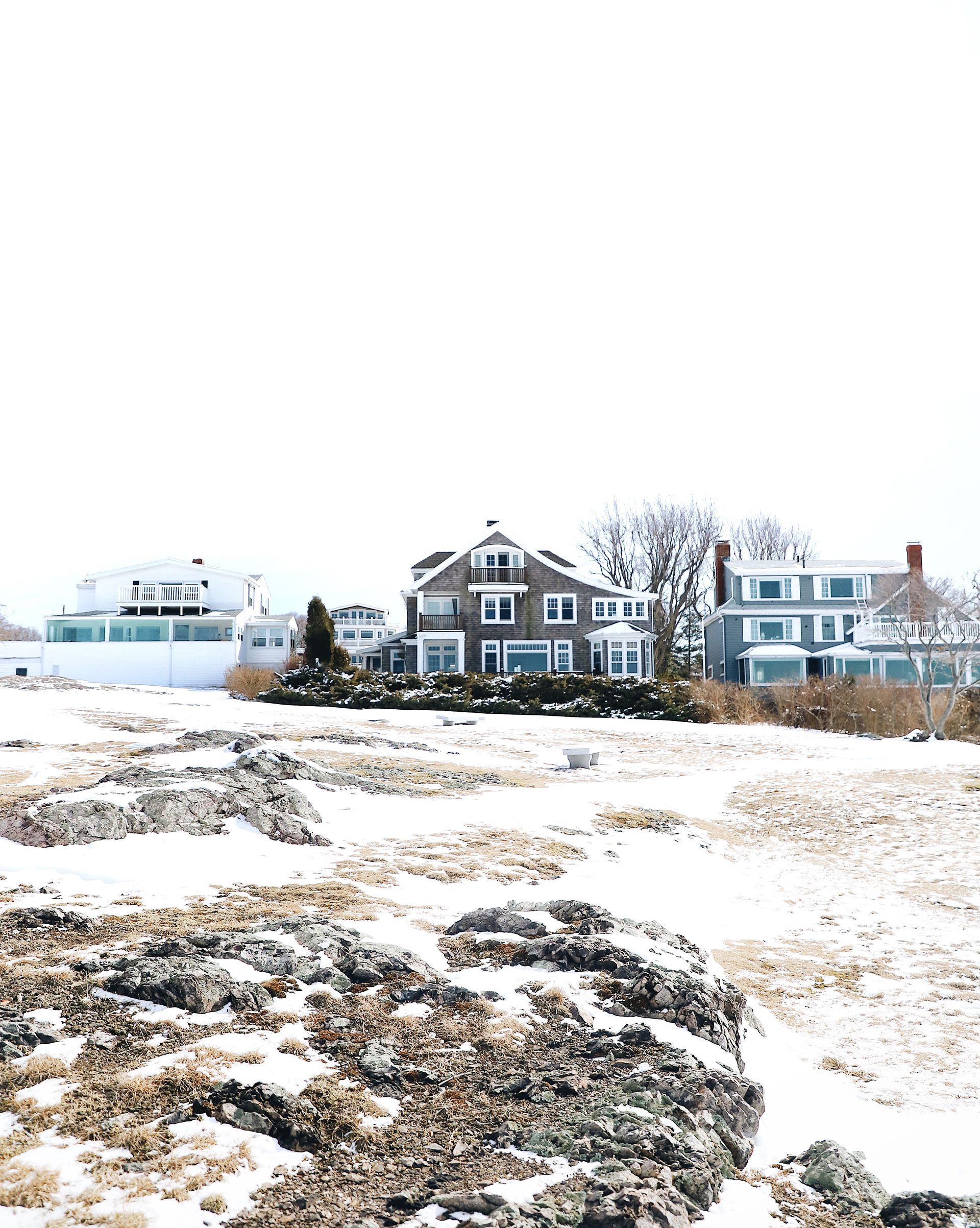 Houses along a rocky, snow-covered coastline under a bright, overcast sky.