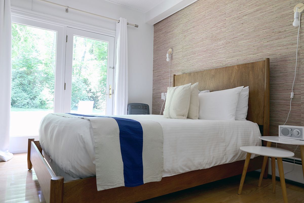 A bright bedroom with a wooden bed, white bedding with a blue runner, and a textured accent wall next to a glass door.