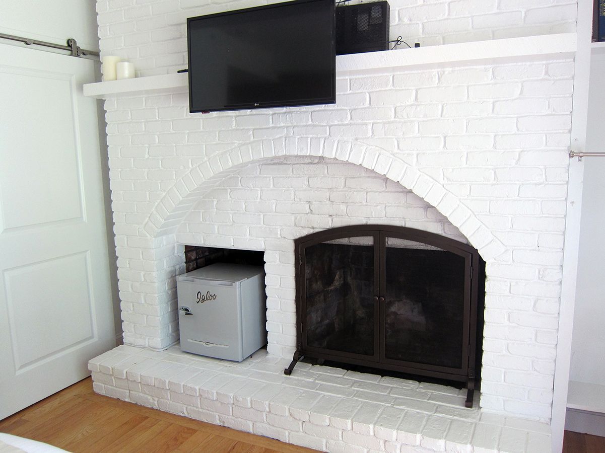 A white brick fireplace with an arched opening containing a metal fireplace screen and a small gray safe inside.