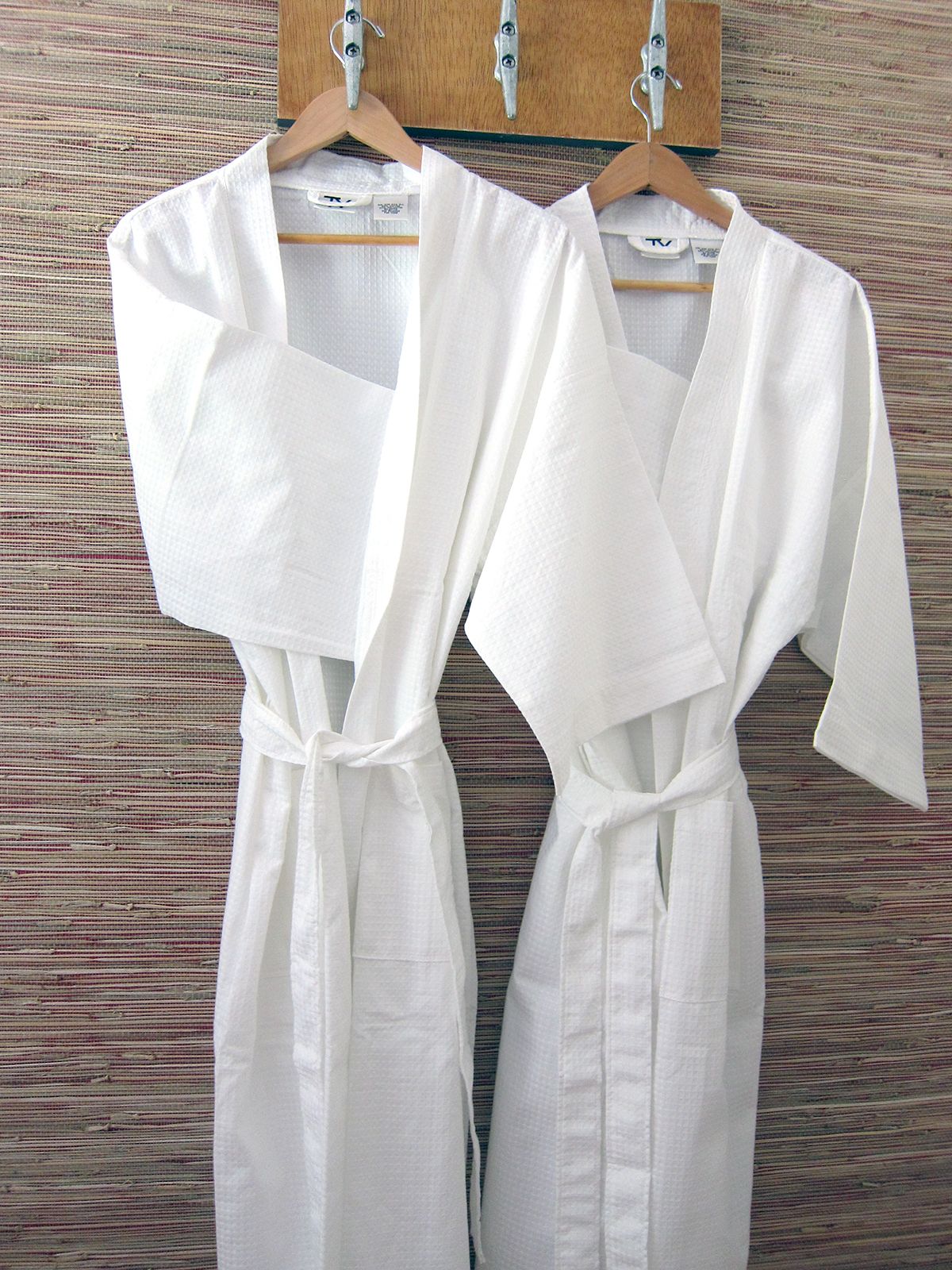 Two clean, white bathrobes hanging on wooden hangers from a hook rack against a textured, light-brown woven wall.
