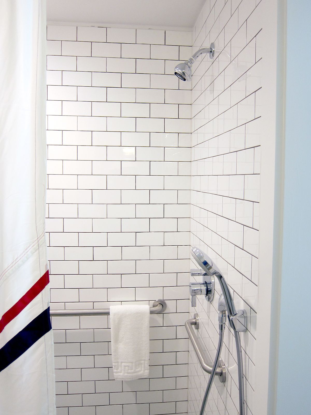 A shower stall featuring white subway tile, a stainless steel shower head, and mounted horizontal grab bars.