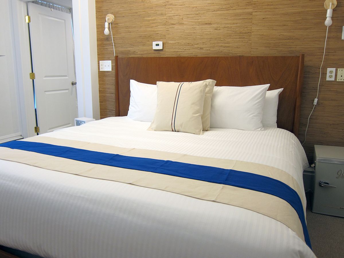 A hotel bed with a tan and blue runner, white pillows, a wood headboard, and wood-paneled walls in a bedroom.