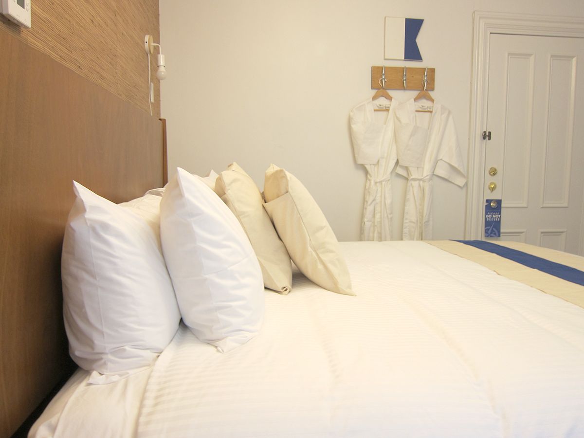 A view of a hotel bed with stacked pillows, next to two hanging white bathrobes on a rack near a closed white door.