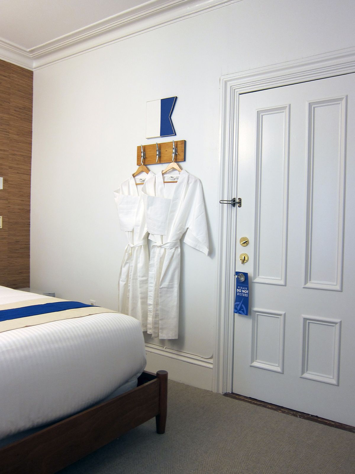 A hotel room features a bed, two white bathrobes hanging on a wall hook, and a white door with a blue sign hanging on it.