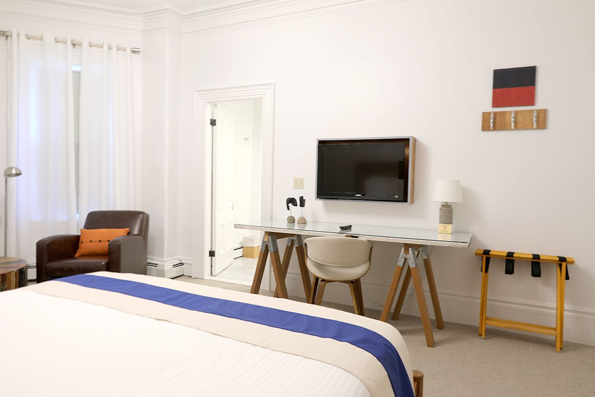 A modern hotel room with a bed featuring a blue runner, a desk with a chair, a wall-mounted TV, and a leather armchair.