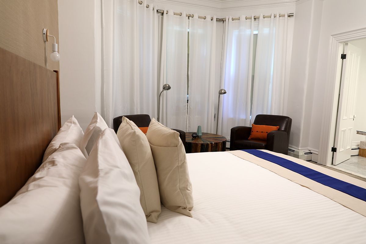 A hotel room featuring a bed with white pillows and a blue runner, a seating area with two dark armchairs, and bay windows.